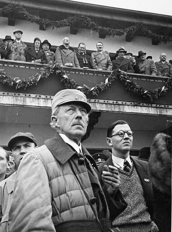 Wilhelm Paulcke (im Vordergrund mit Mütze) bei der Eröffnungsfeier der Olympischen Winterspiele in Garmisch-Partenkirchen, im Hintergrund Adolf Hitler, Joseph Goebels und Hermann Göring, 6. Februar 1936